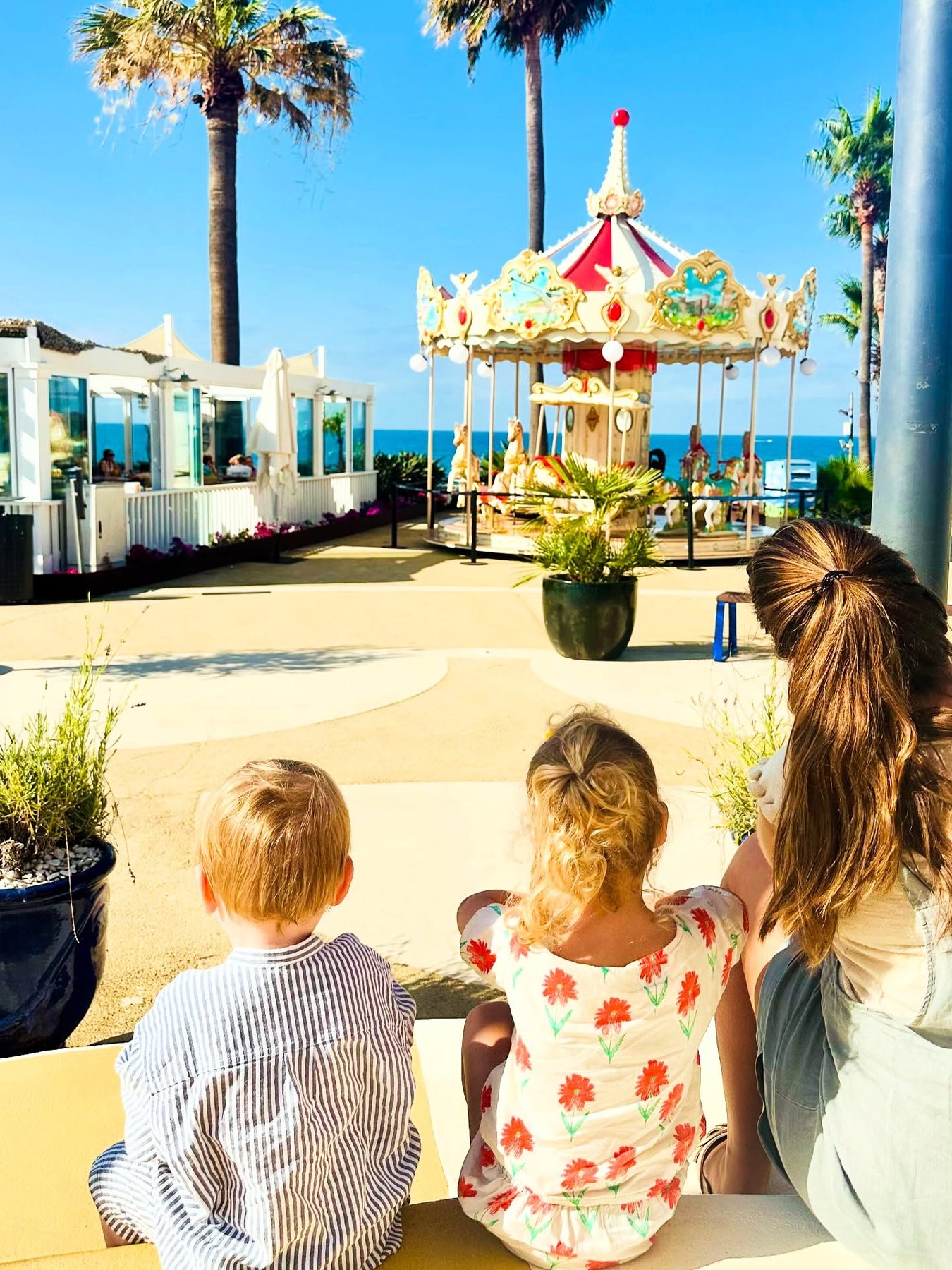 A FunWorks babysitter sitting with two children on a seafront promenade in the Algarve, watching a carousel beside the sea and palm trees