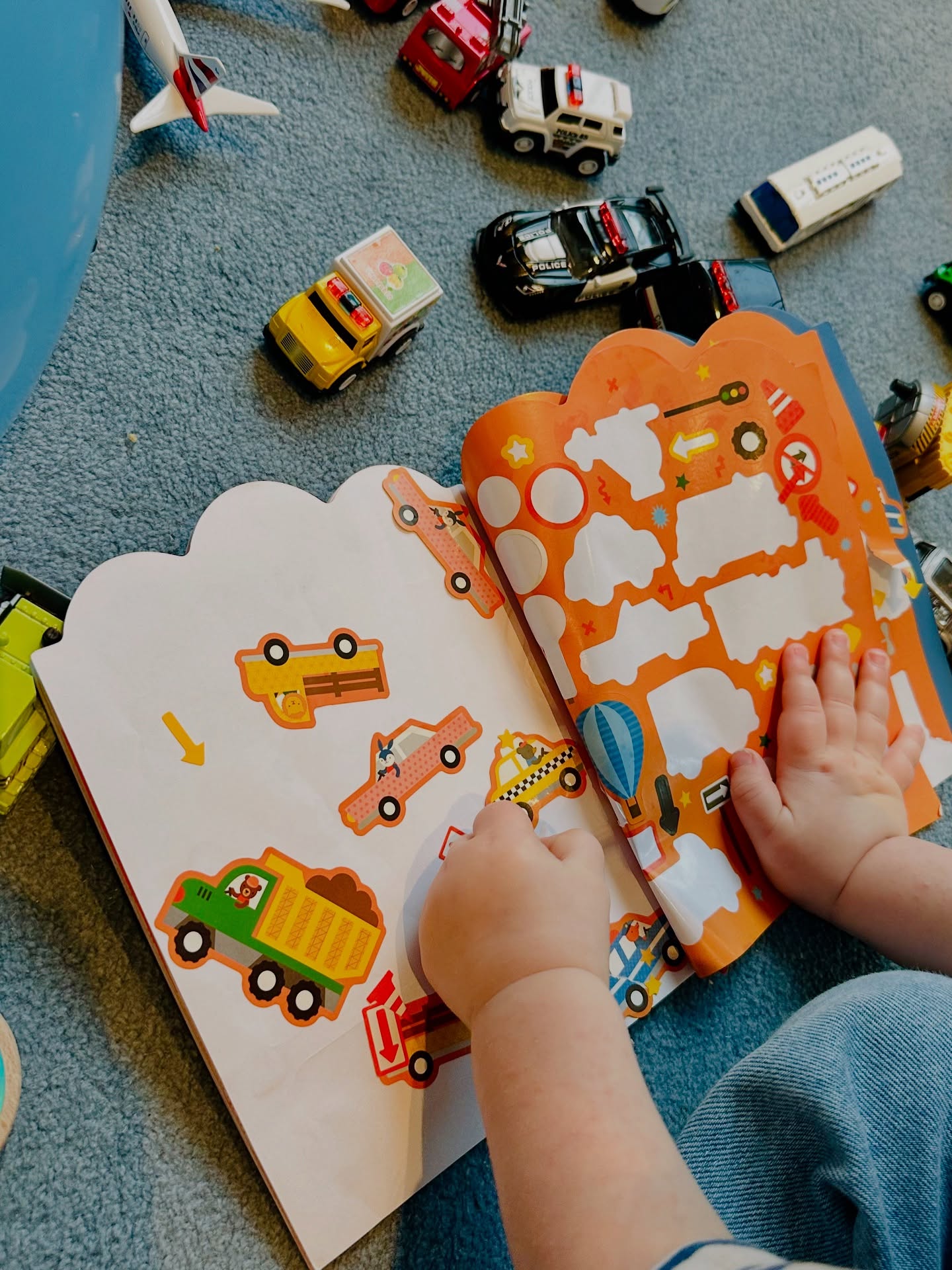 A child's hands working through a vehicle sticker book, surrounded by toy cars on a carpet