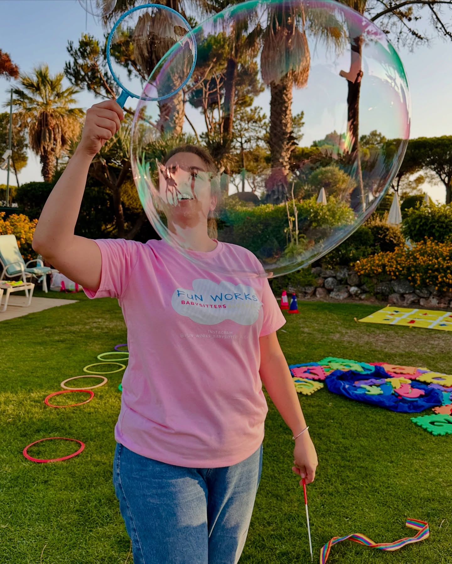 A FunWorks babysitter in a branded pink T-shirt making a giant soap bubble in a sunlit Algarve garden