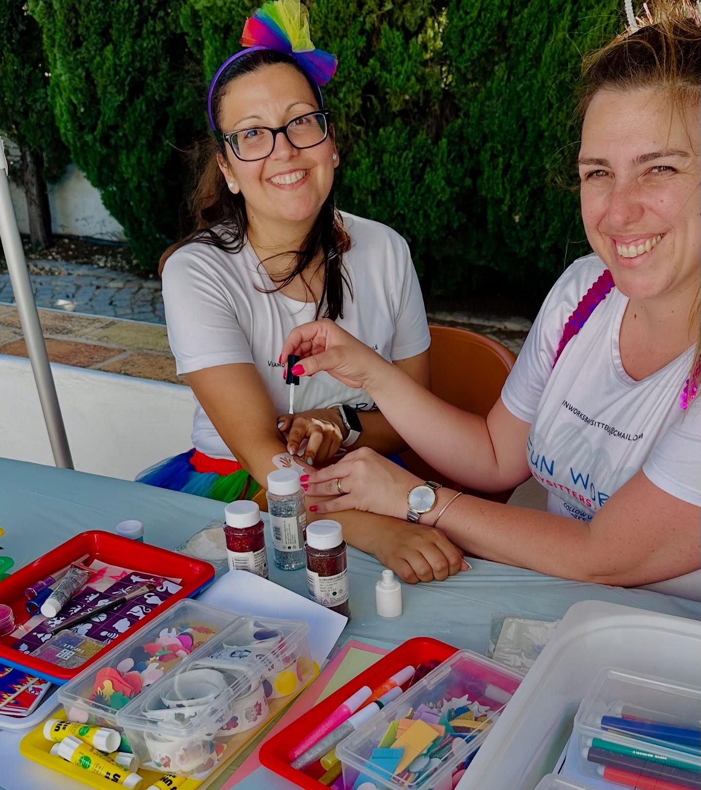 Two FunWorks babysitters in branded T-shirts at an outdoor craft table set up with glitter, stickers and pens