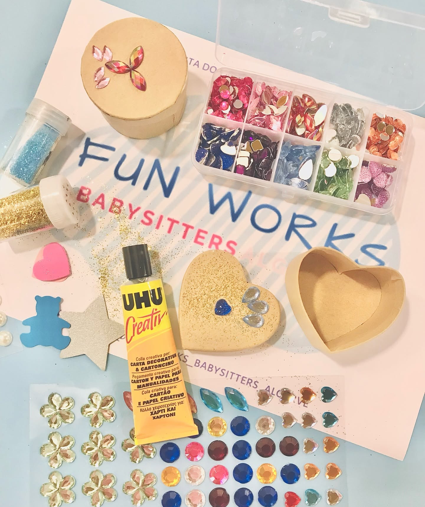 Craft materials laid out on FunWorks Babysitters branded paper — glitter, gems, glue, paper hearts and stars